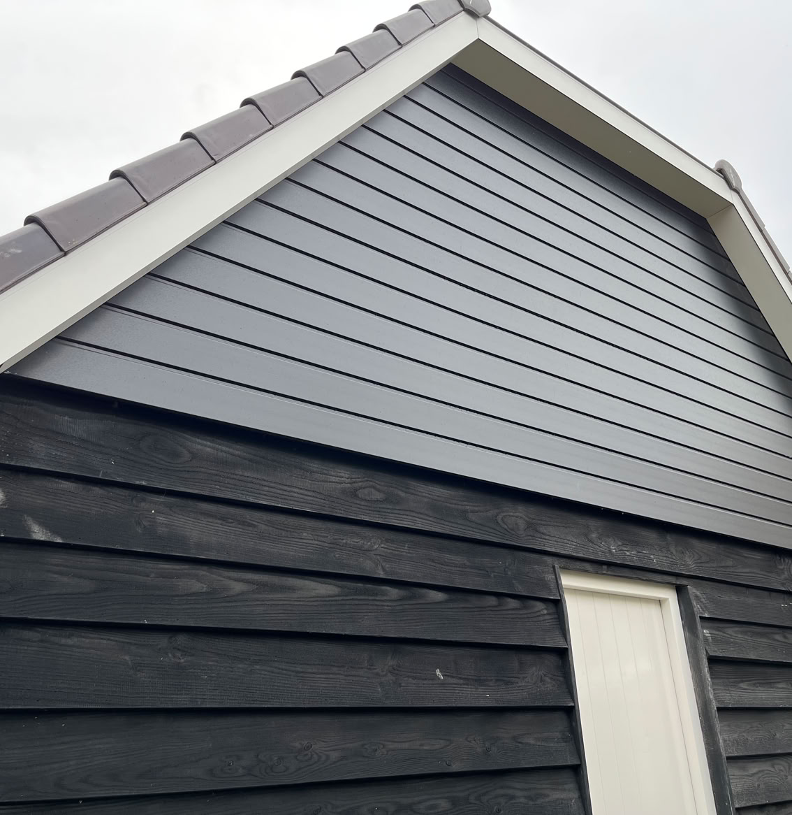 Zijkant van een gebouw met zwarte houten gevelbekleding en een witte deur, onder een grijs zadeldak.