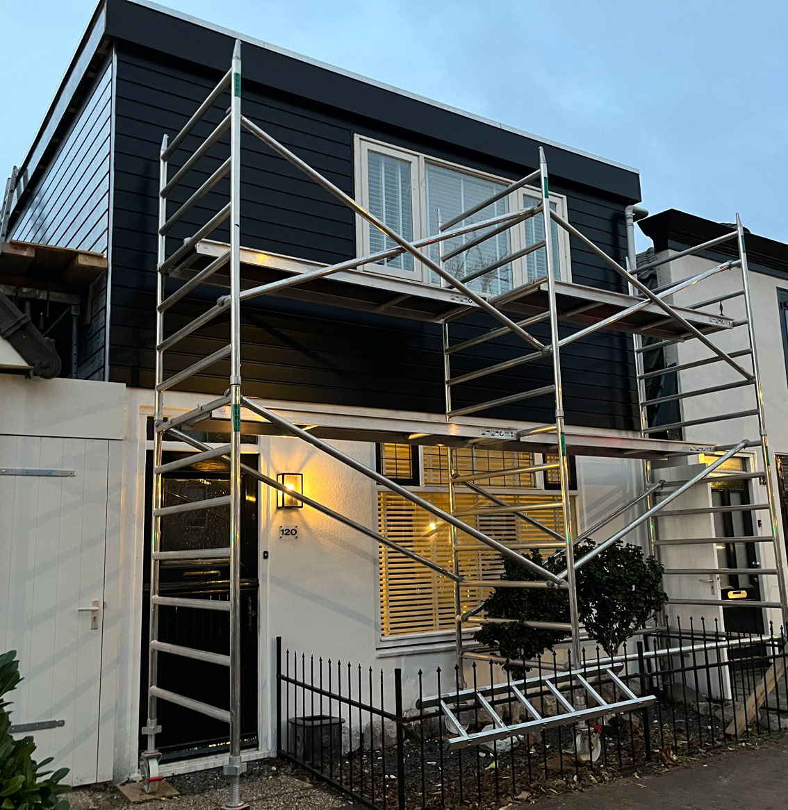 Een huis met een steiger eromheen, verlichting aan de voorkant, en een ladder op de grond.