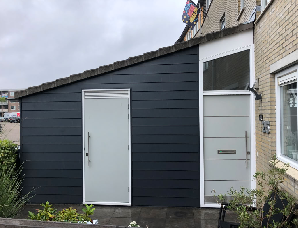 Donkergrijze aanbouw met een witte deur en een raam, naast een bakstenen gebouw.